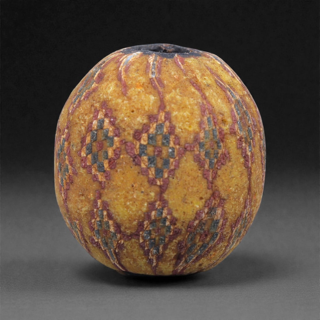 Ancient Egypto-Roman Mosaic Glass Bead - Yellow Ground Checkerboard Sphere, 84J: Formed in opaque yellow mosaic glass, this spherical bead is decorated with repeating vertical registers of checkerboard cane clusters composed of red, green, blue, and cream tesserae. The structured 
