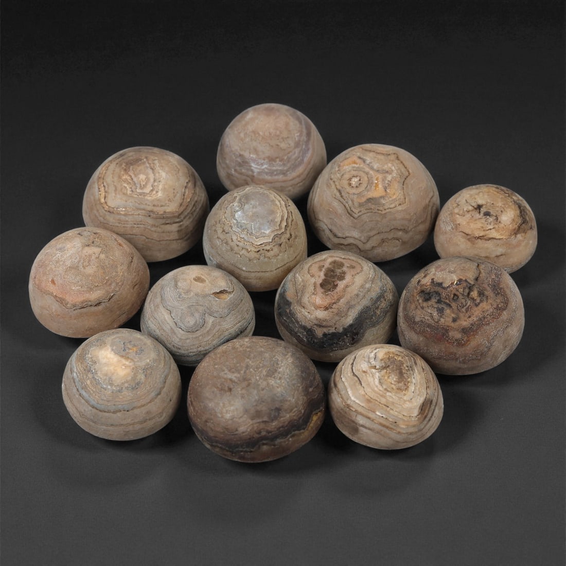 Tibetan Stone Luk Mik Beads, Possibly 19th-20th Century, 57LE: Worked from banded stone with natural concentric veining, the group comprises twelve hemispherical bead forms, each shaped with a rounded dome and a flattened base. The surfaces show layered mineral a