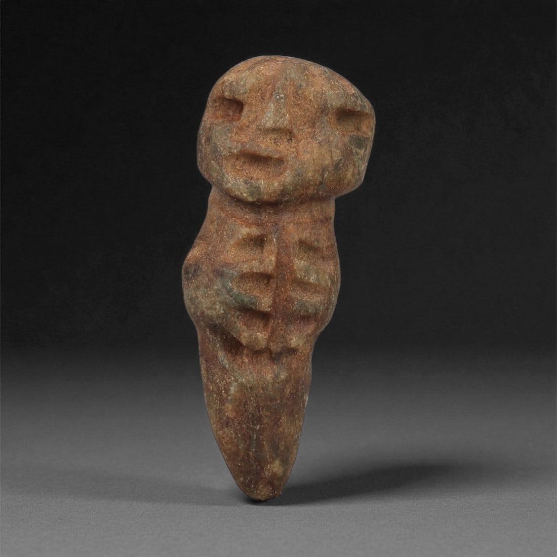 A Taino Carved Stone Idol, Late Pre-Columbian Period, Circa 1000-1500 CE, 91LF: Carved from a mottled brown-green stone with natural surface inclusions, the figure presents an elongated tapering lower body terminating in a pointed base. The torso is defined by a series of incised
