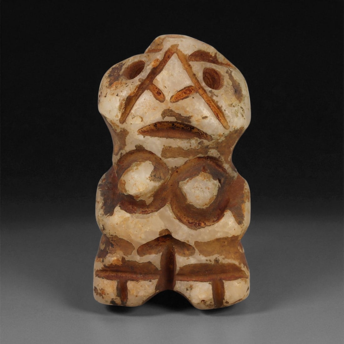 A Taino Carved Stone Idol, Late Pre-Columbian Period, Circa 1000-1500 CE, 11ZB: Carved from mottled cream and brown stone, the figure stands with short legs and a compact torso. The head is modeled with recessed circular eyes, a sharply triangular nose, and a shallow incised mout