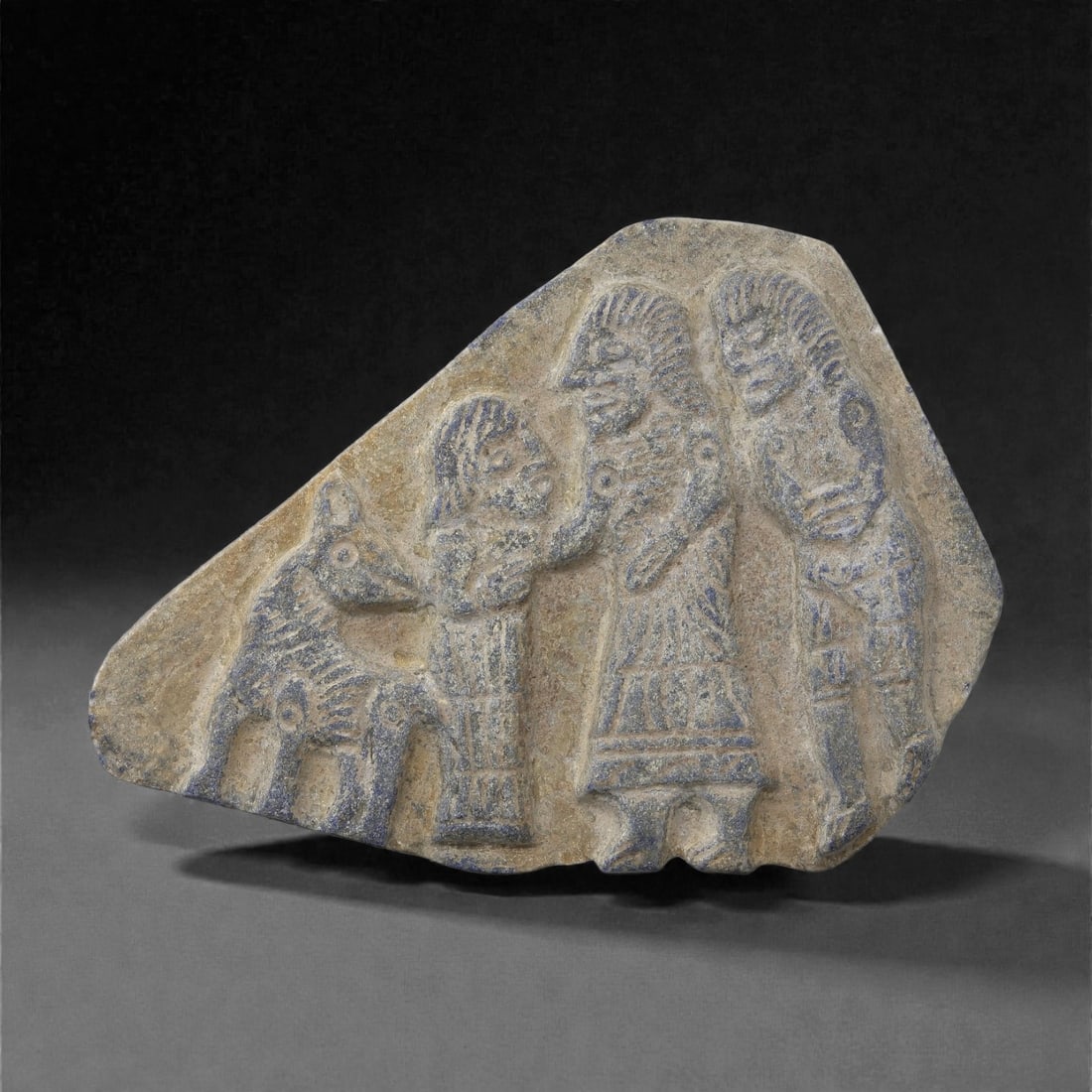 A Kassite Lapis Lazuli Plaque Relief, 11LM: Carved from a blue-grey lapis lazuli slab with natural veining, the plaque is worked in shallow relief with three standing figures and a small quadruped arranged across the surface. The figures are sh