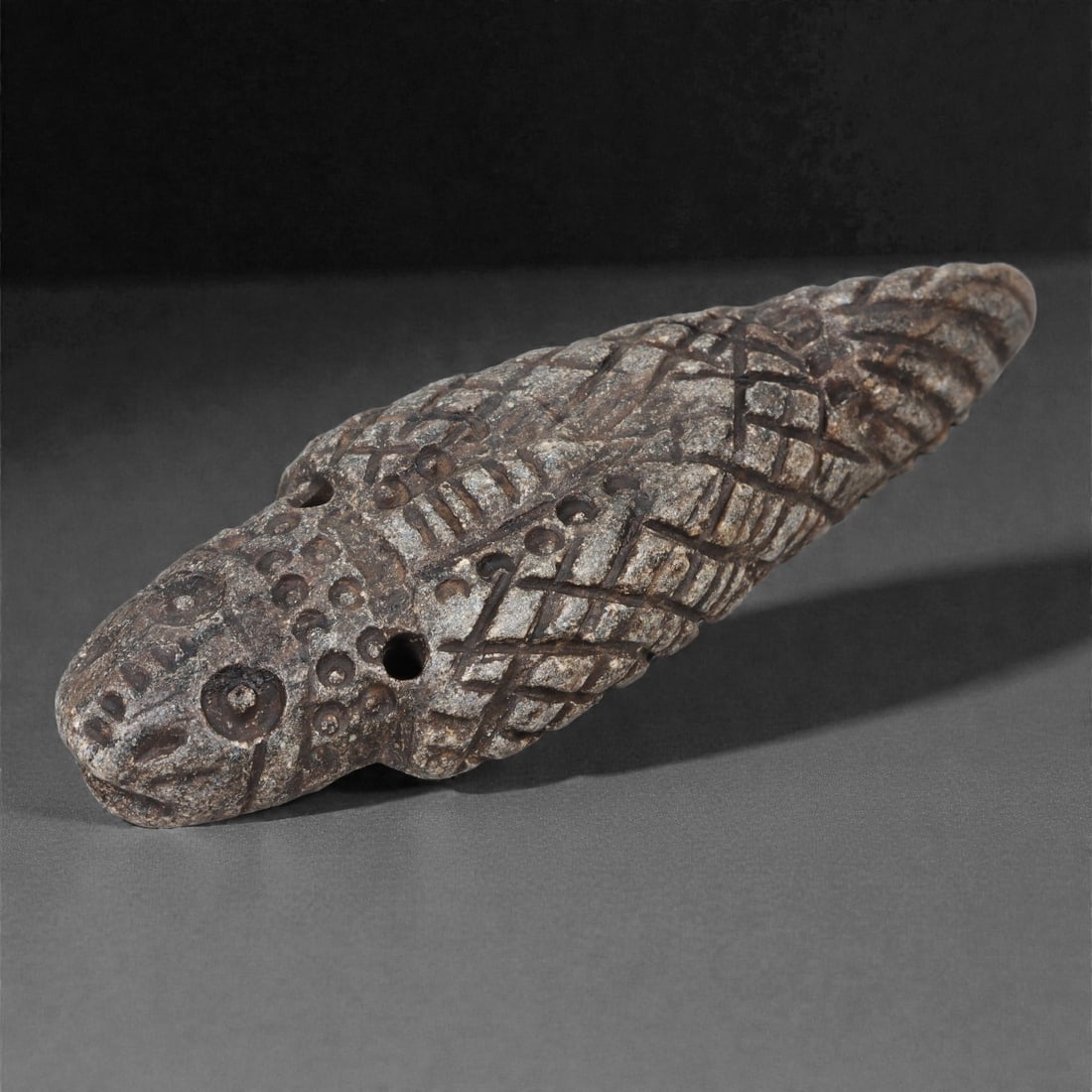 A Syro-Anatolian Schist Zoomorphic Amulet, Iron Age, 1st Millennium BCE, 55LJ: Carved from dark grey schist, the amulet is shaped as an elongated reptilian creature with a rounded head, recessed circular eyes, and a short muzzle. The body tapers toward the tail and is covered wi