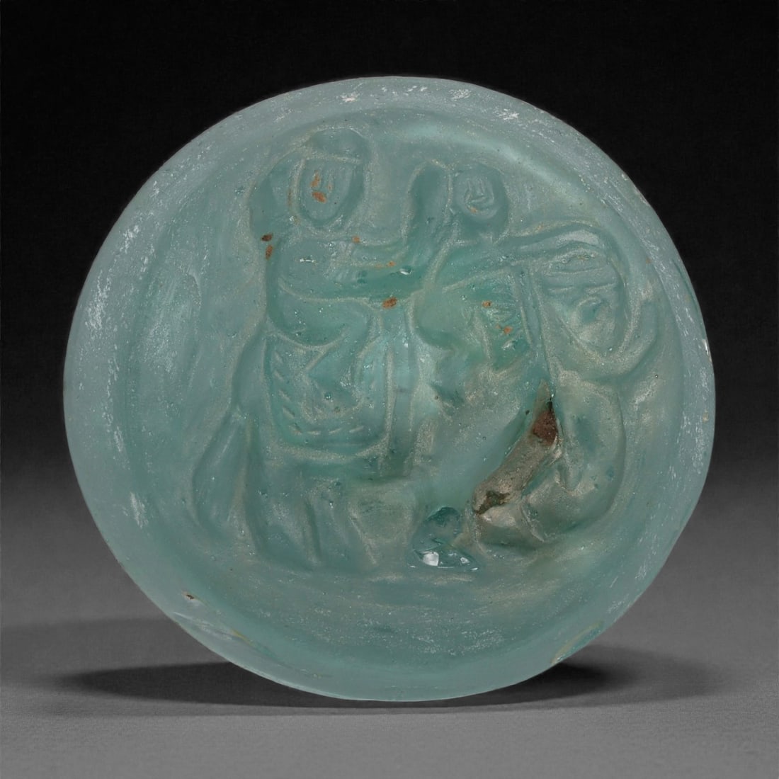A Sasanian Figures Riding an Elephant Glass Medallion, 3rd-7th Century CE, 29LP: Cast from translucent blue-green glass, the medallion is circular in form and decorated in low molded relief with a scene of two human figures positioned atop an elephant. The riders are shown seated