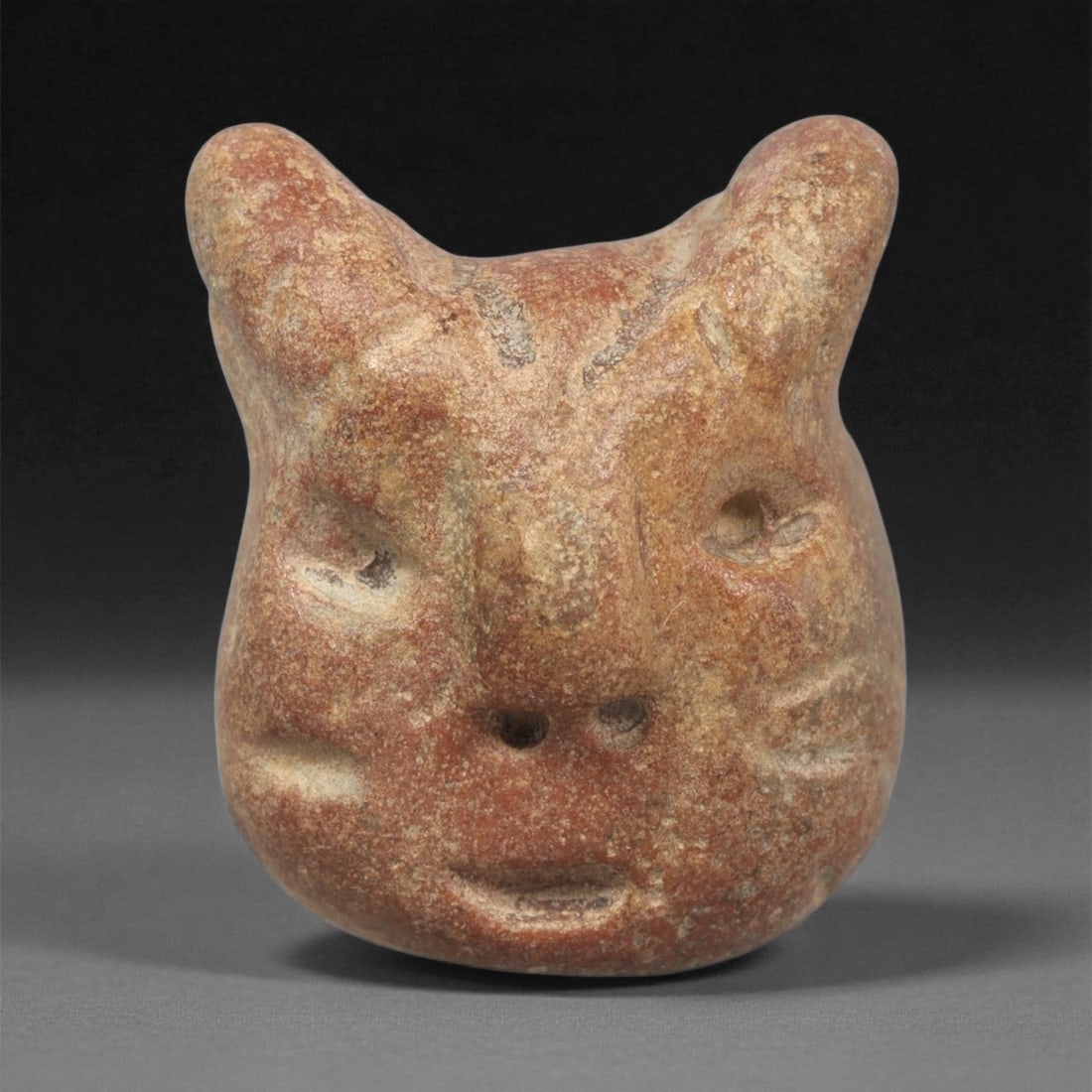 A Mediterranean Stone Maskette, 12th-17th Century, 19LC: Carved from a reddish-brown stone with lighter surface mottling, the maskette presents a rounded form surmounted by two prominent projections resembling stylized ears or horn-like extensions. The faci