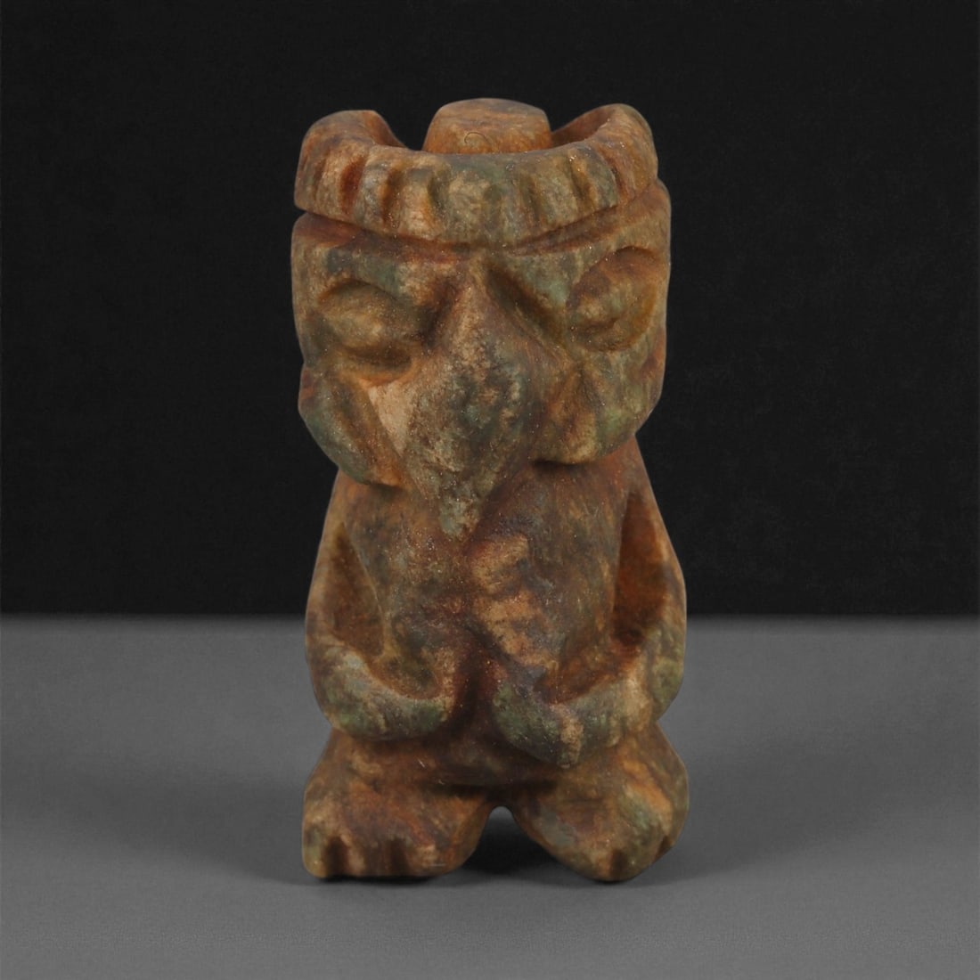 A Taino Carved Stone Idol, Late Pre-Columbian Period, Circa 1000-1500 CE, 67ZA: Carved from mottled brown and green stone, the figure is modeled in a seated posture with bent legs and arms resting on the abdomen. The head is defined by a pronounced brow band, deeply recessed eyes