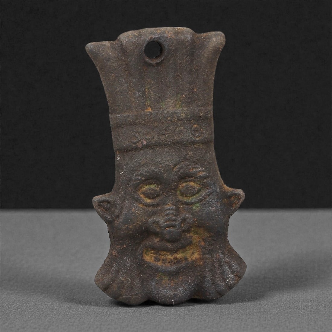An Egyptian Bronze Plaque of Bes, Late Period to Ptolemaic, 7th-1st Century BCE, 52LP: Cast in bronze, the plaque depicts the dwarf-protective deity Bes shown frontally with characteristic leonine facial features, rounded eyes, and a protruding tongue. The figure wears a tall crown with