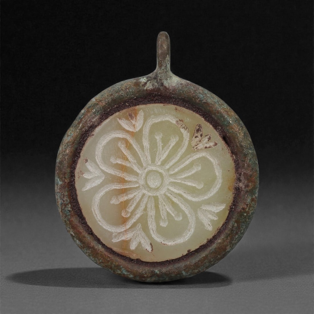 An Islamic Bronze Amulet with Agate Rosette Inset, 10th-13th Century CE, 13LP: Cast in bronze with a circular form and an integral suspension loop, the amulet encloses a fitted disc of pale agate carved in low relief with a rosette composed of six rounded petals encircling a cen