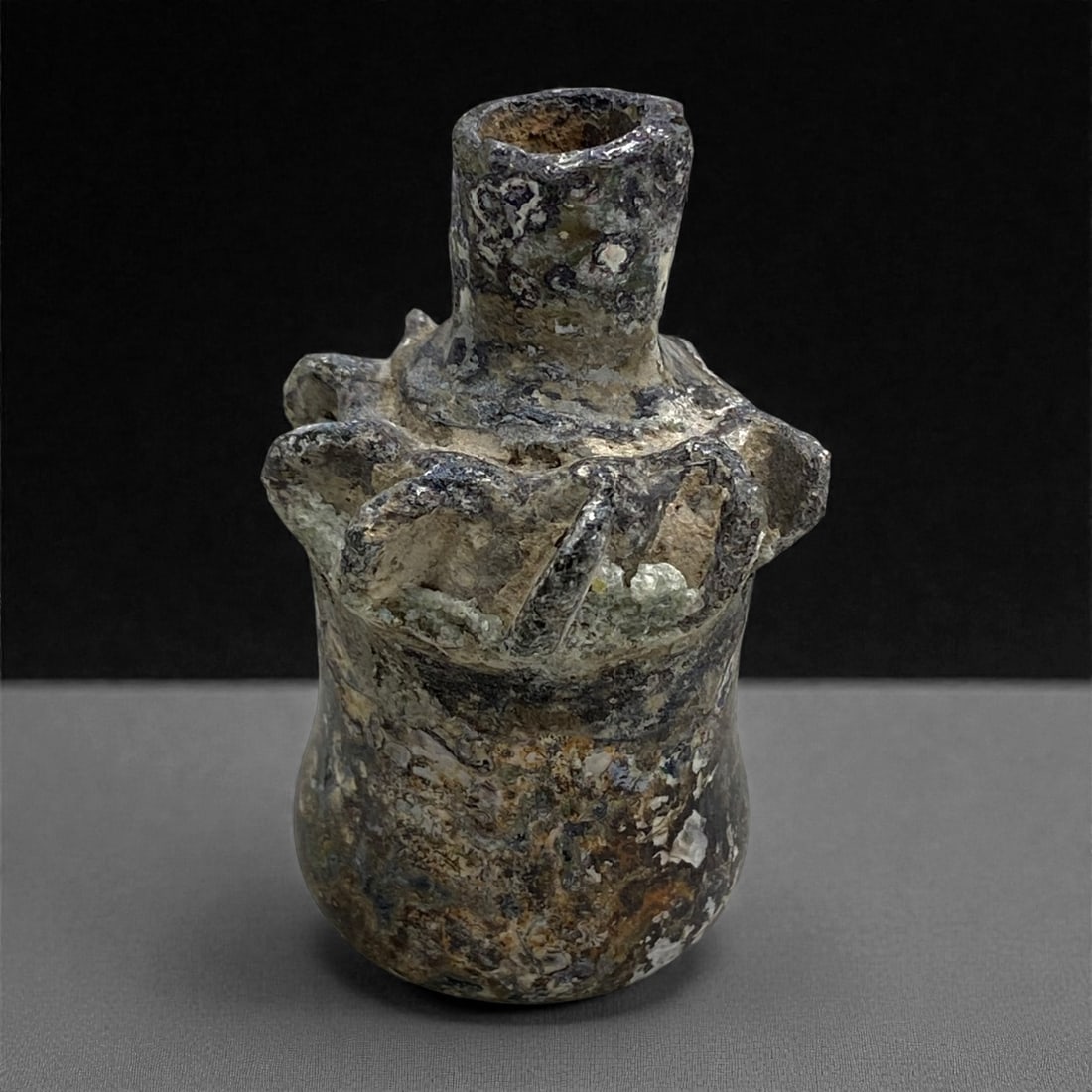A Roman Levantine Glass Unguentarium with Pinched Collar, 1st Century CE, 42 g, 30AA: Blown from pale green glass now obscured by dense weathering and mineral accretion, the vessel is formed with a narrow cylindrical neck above a rounded body, the shoulder shaped with a series of deepl