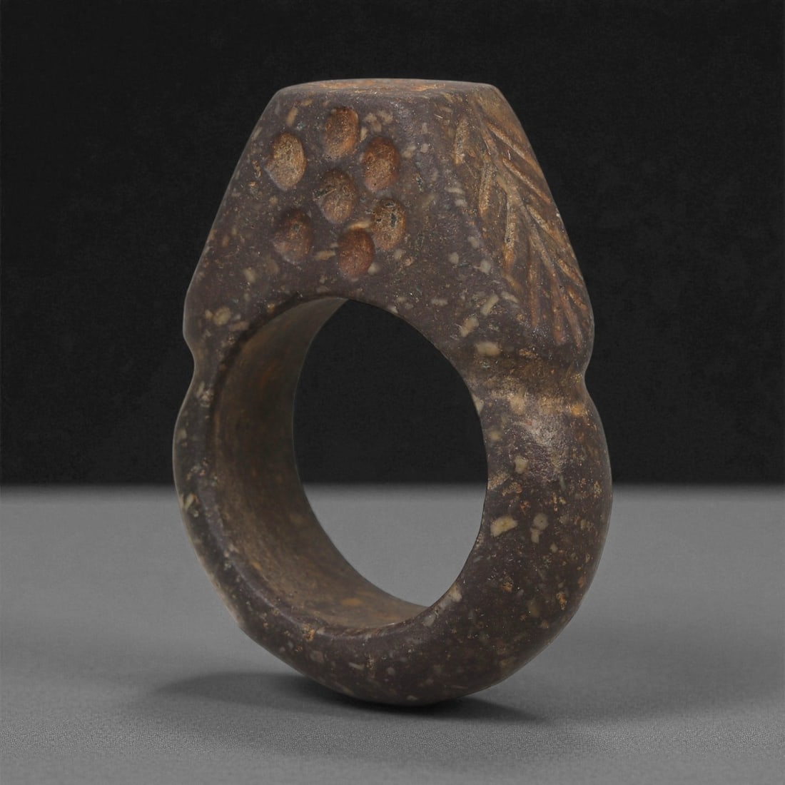 An Indo-Parthian Stone Ring, 1st-3rd Century CE, 15LD: Carved from a dark stone with lighter mineral inclusions, the object is shaped as a thick circular ring with a flattened exterior panel. The outer surface bears incised linear motifs forming a chevron