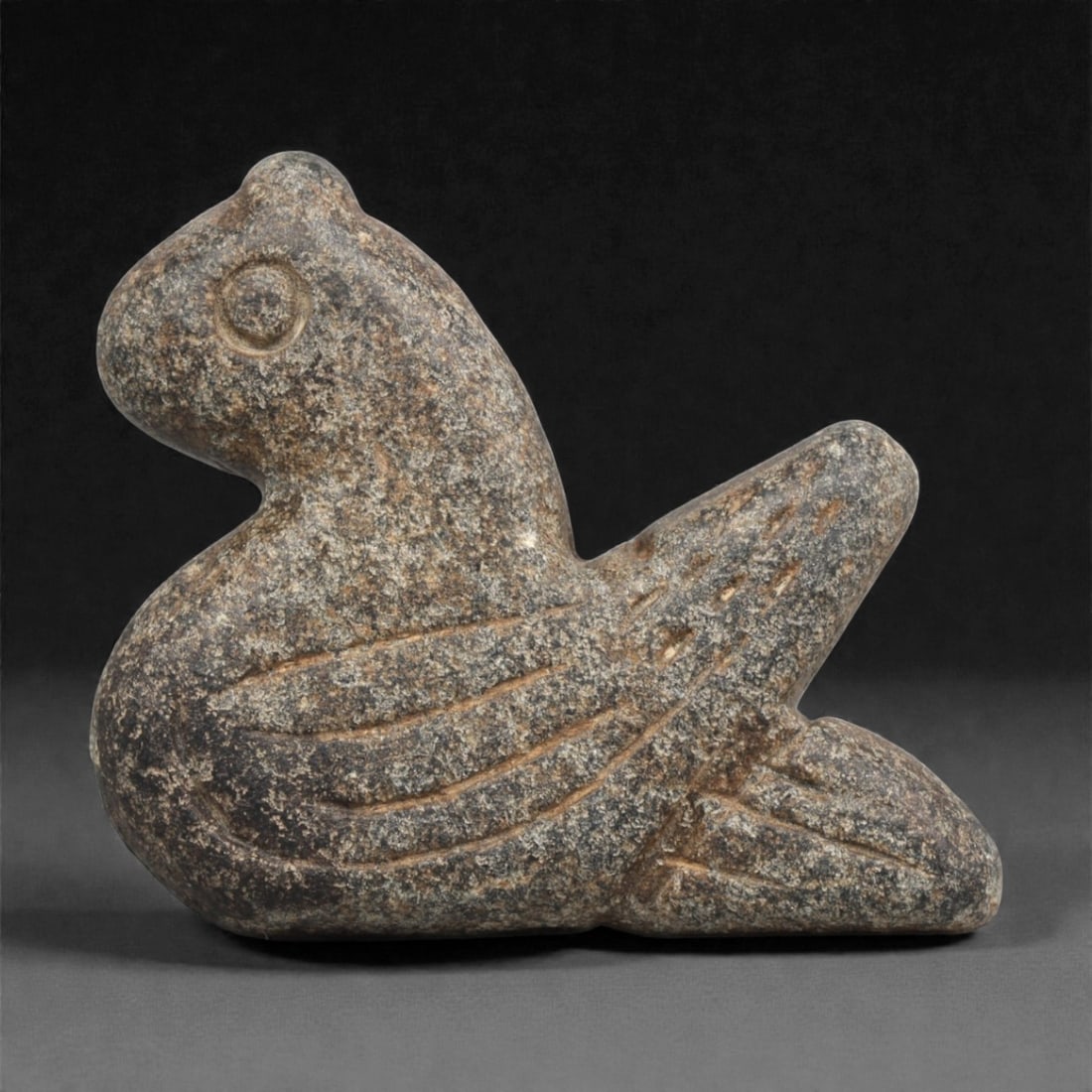 A Near Eastern Stone Bird Amulet, 3rd-2nd Millennium BCE, 75ZA: Carved from a mottled dark stone with granular inclusions, the object is shaped as a stylized bird in profile with a rounded head and a short beak. The body is compact, with incised linear grooves del
