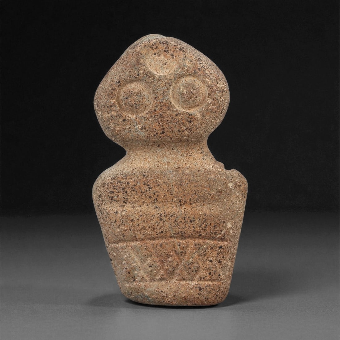 A Near Eastern Stone Anthropomorphic Idol, 4th-3rd Millennium BCE, 52ZA: Carved from a light brown stone with dense dark inclusions, the figure is shaped with a rounded head bearing two recessed circular eyes defined by concentric grooves and a small circular depression ab