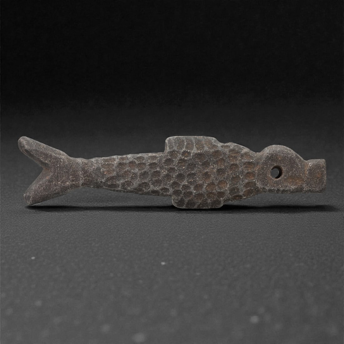 A Mesopotamian Steatite Fish Amulet, Iron Age, 1st Millennium BCE, 85LR: Carved from dark grey steatite, the amulet is shaped as an elongated fish with a bifurcated tail and a flattened body. The surface is decorated with a regular pattern of shallow circular depressions s