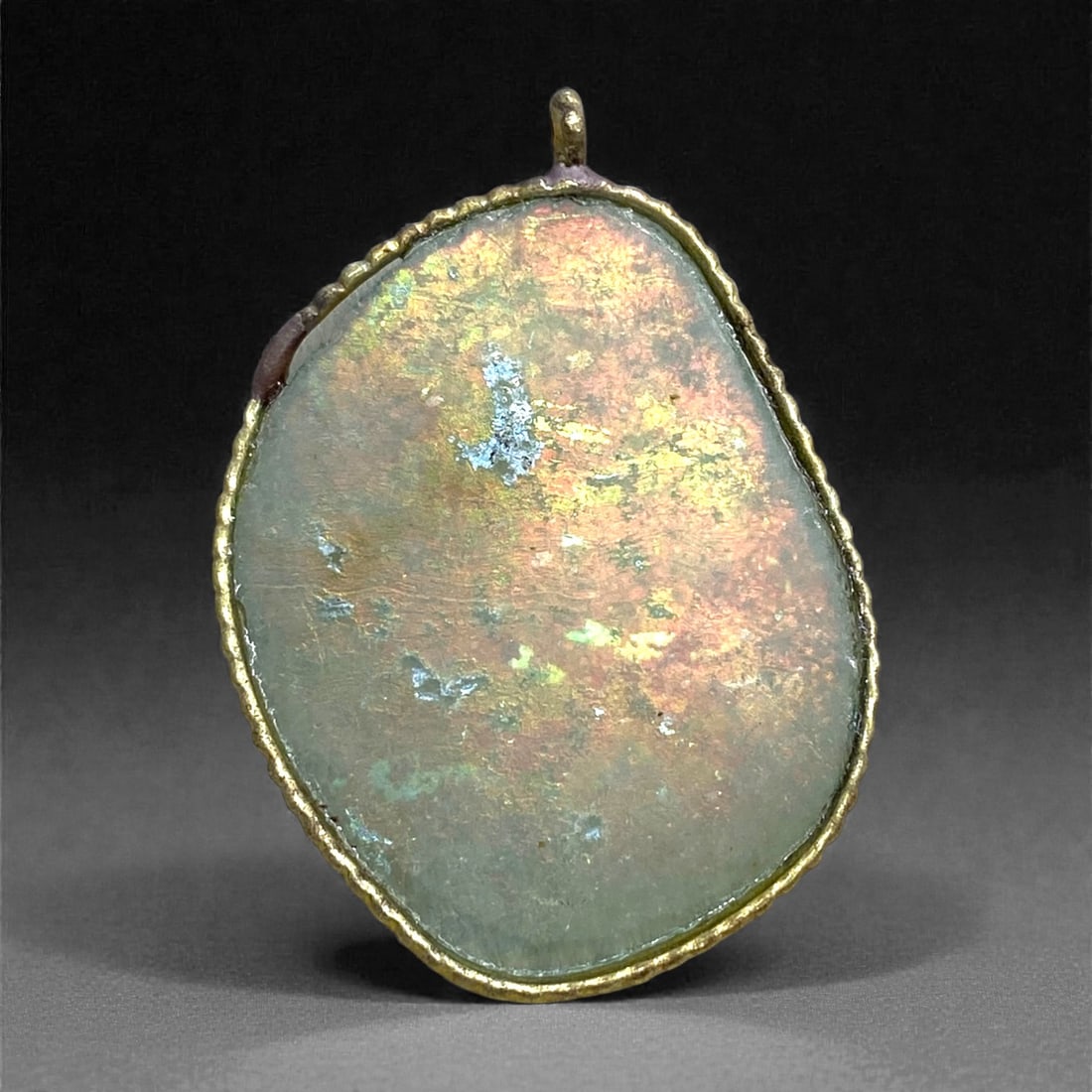 A Roman Glass Pendant, the Glass 1st-4th Century CE, Mounted in a Modern Setting, 69AA: Composed of a thin fragment of ancient Roman glass exhibiting a soft green tone beneath broad areas of iridescent weathering in gold, pink, and pale blue, the surface shows the smooth, laminar degrada