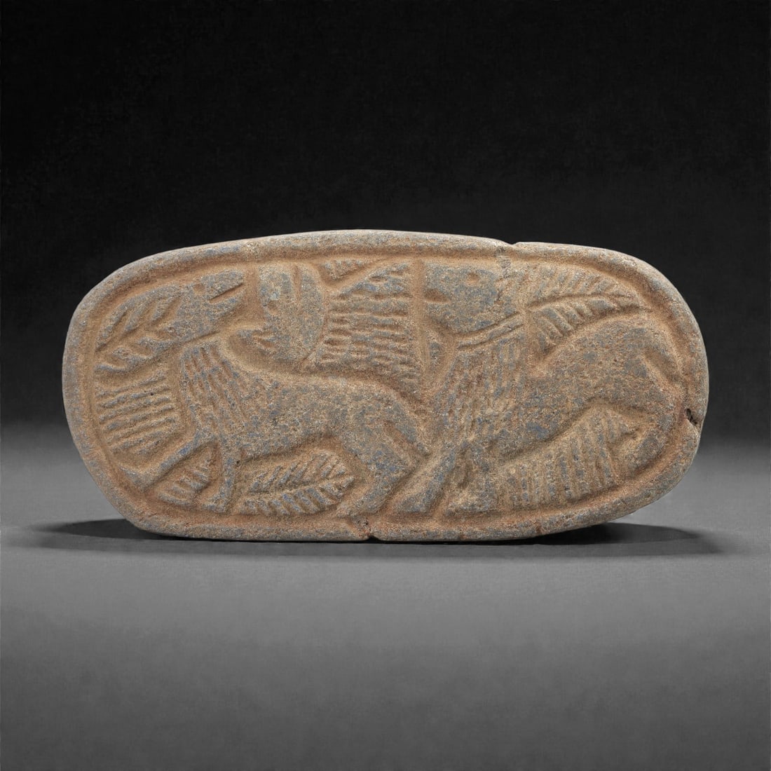 A Sasanian Lapis Lazuli Oval Plaque with Confronted Quadrupeds, 3rd-7th Century CE, 89LQ: Carved from lapis lazuli with brown archaeological surface accretions, the elongated oval plaque is worked in shallow relief depicting two confronted quadrupeds arranged symmetrically around a central