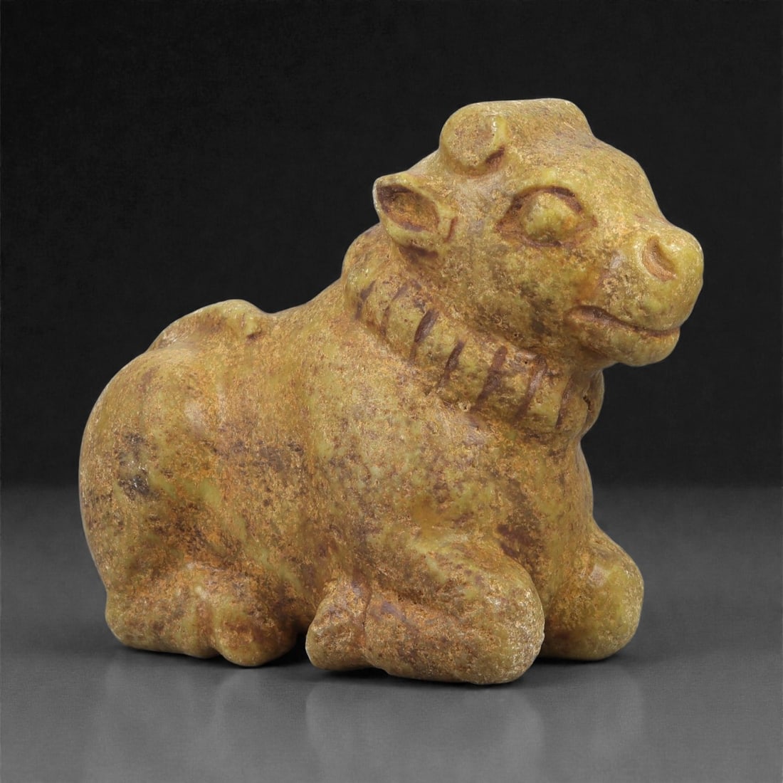 An Achaemenid Serpentine Bovid Figurine, 13LM: Carved from yellow-brown serpentine with scattered surface patination, the figurine depicts a recumbent bovid with the forelegs folded beneath the chest and the hindquarters compactly tucked under the