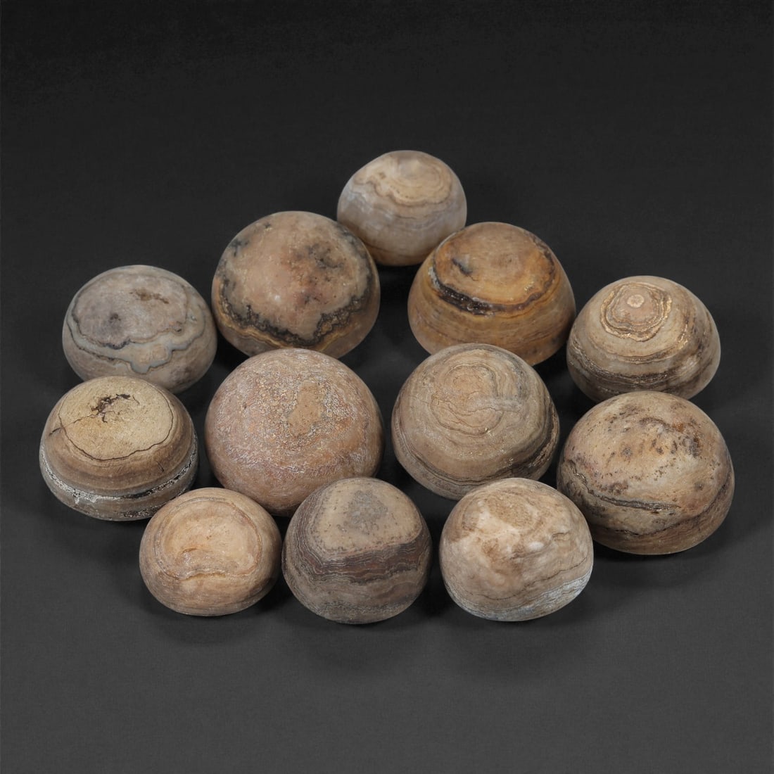 Tibetan Stone Luk Mik Beads, Possibly 19th-20th Century, 59LE: Worked from banded stone with natural concentric veining, the group comprises twelve hemispherical bead forms, each shaped with a rounded dome and a flattened base. The surfaces show layered mineral a