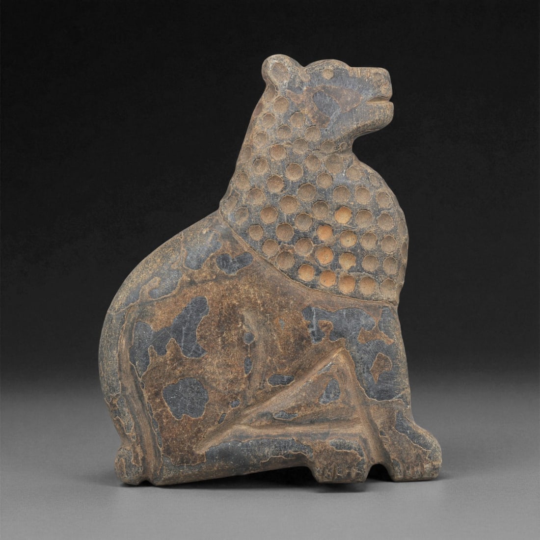An Indo-Parthian Stone Lion Plaque, 1st-3rd Century CE, 35ZA: Carved from a dark stone with lighter surface accretions, the plaque depicts a seated lion in profile. The head and upper body are covered with a field of circular depressions, while the lower body be