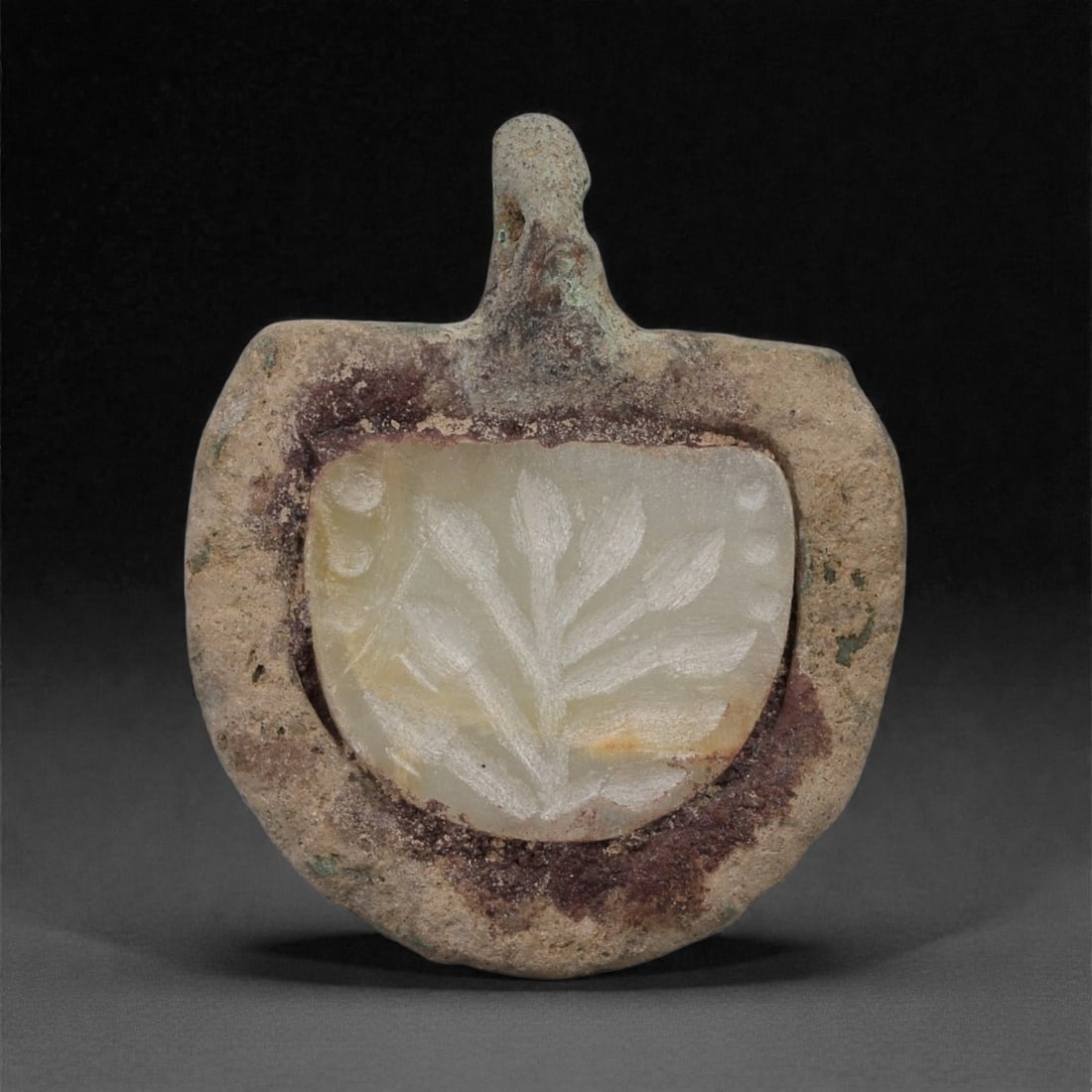 An Islamic Bronze Amulet with Agate Inset, 10th-13th Century CE, 14LP: Cast in bronze and formed with a rounded outline and an integral suspension loop, the amulet encloses a fitted agate plaque carved in low relief with a stylized vegetal motif consisting of a central s