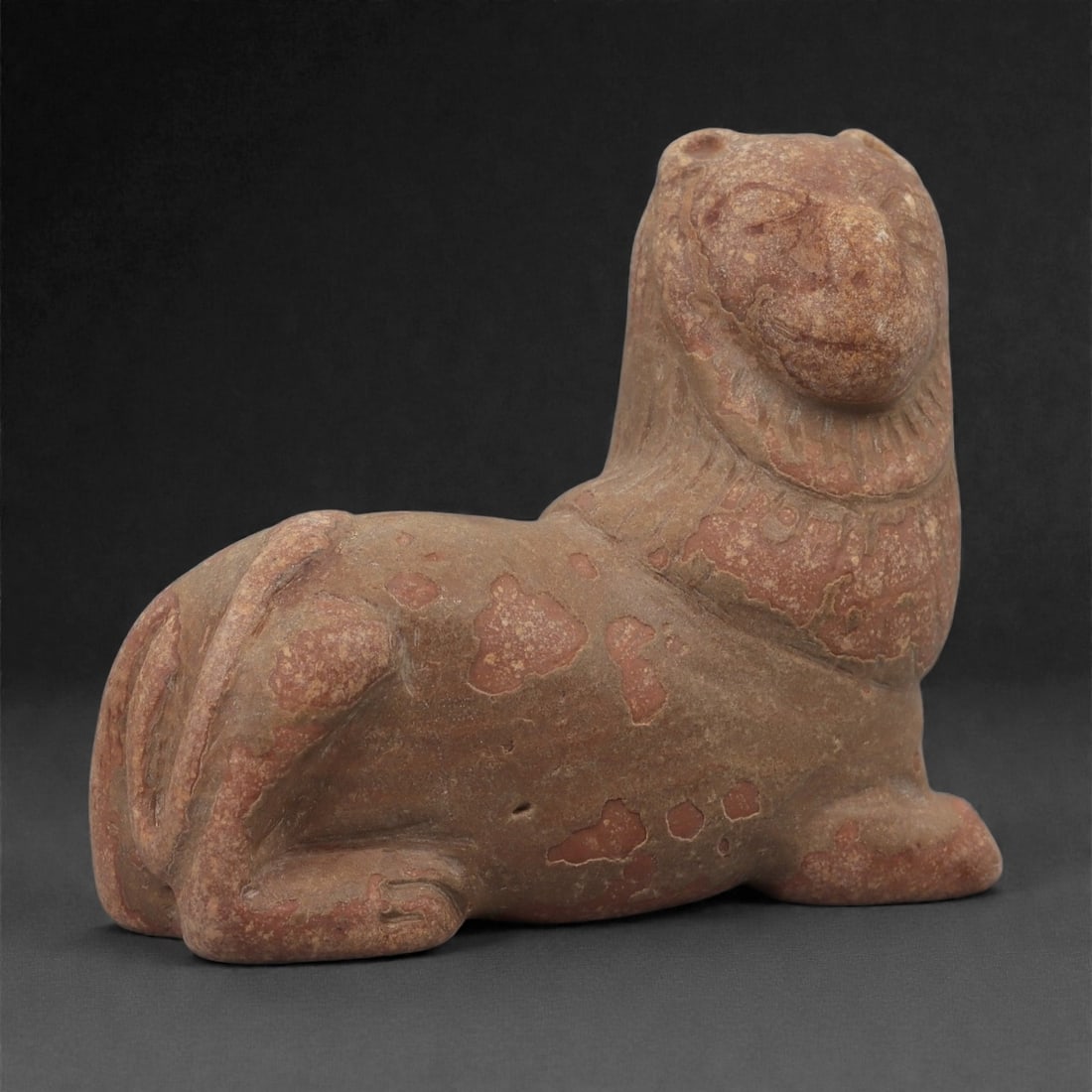 An Anatolian Iron Age Stone Lion Effigy, Circa 9th-7th Century BCE, 30LQ: Carved from reddish stone with natural surface variations, the figure represents a recumbent lion with the forelegs folded beneath the body and the hindquarters set compactly to the side. The head is