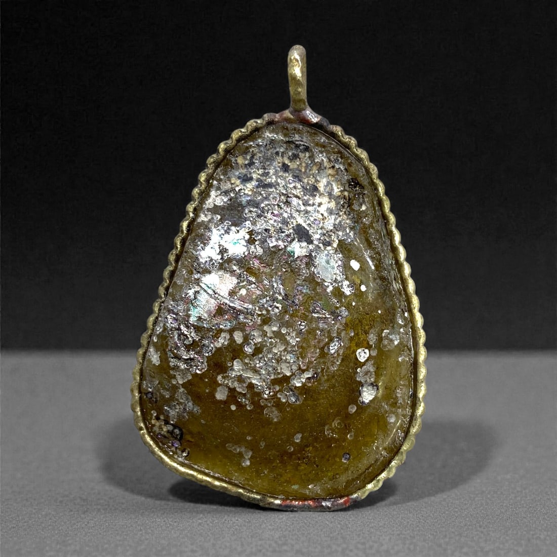 A Roman Glass Pendant, the Glass 1st-4th Century CE, Mounted in a Modern Setting, 64AA: Made from a fragment of ancient Roman glass retaining a translucent brown tone beneath areas of encrustation and iridescent weathering, the surface exhibits granular pitting and patches of laminated s