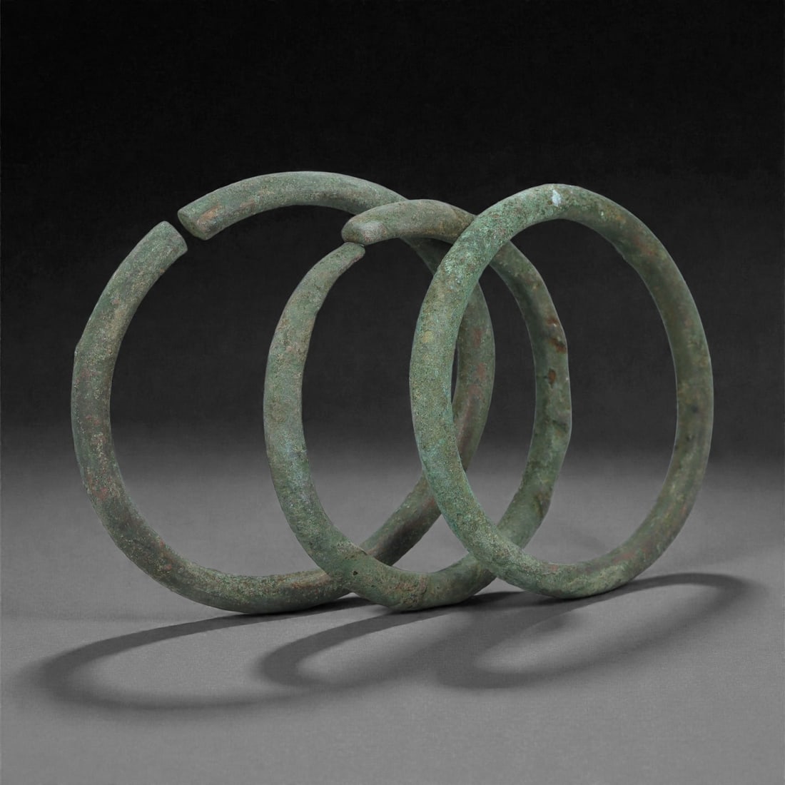 Roman Bronze Bracelets, 1st-3rd Century CE, 2H: Cast in bronze with a mottled green patina, the group comprises three circular bracelets of uniform round-section profile, each terminating in slightly offset ends indicating an open form. The surface
