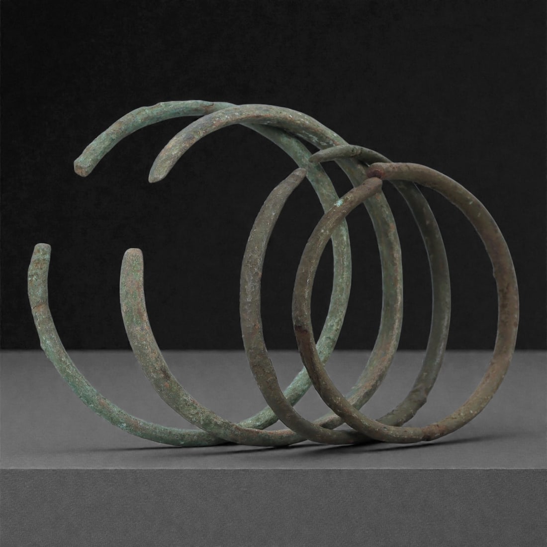 Roman Bronze Bracelets, 1st-3rd Century CE, 12H: Cast in bronze with slender circular profiles, the group comprises four open bracelets exhibiting green and brown patination with areas of surface accretion consistent with burial conditions. Each bra