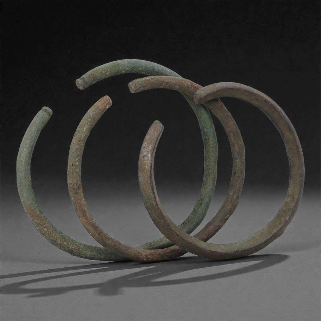 Roman Bronze Bracelets, 1st-3rd Century CE, 19H: Cast in bronze with rounded circular profiles, the group comprises three open bracelets exhibiting green and brown patination with localized surface accretions typical of burial conditions. Each brace