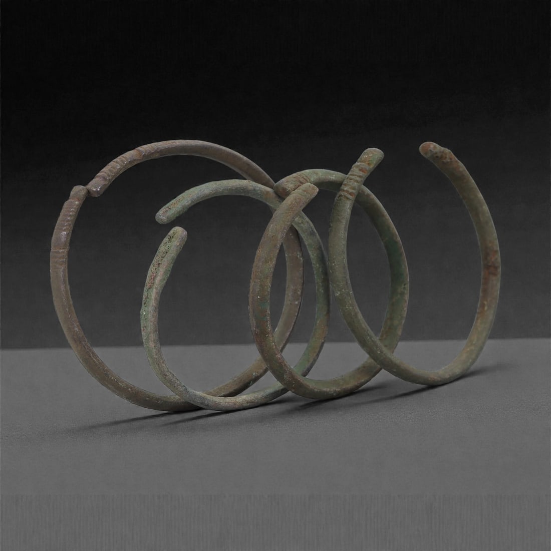 Roman Bronze Bracelets, 1st-3rd Century CE, 8H: Cast in bronze with slender circular profiles, the group comprises four small open bracelets exhibiting green and brown patination with surface accretions consistent with burial conditions. Each brace