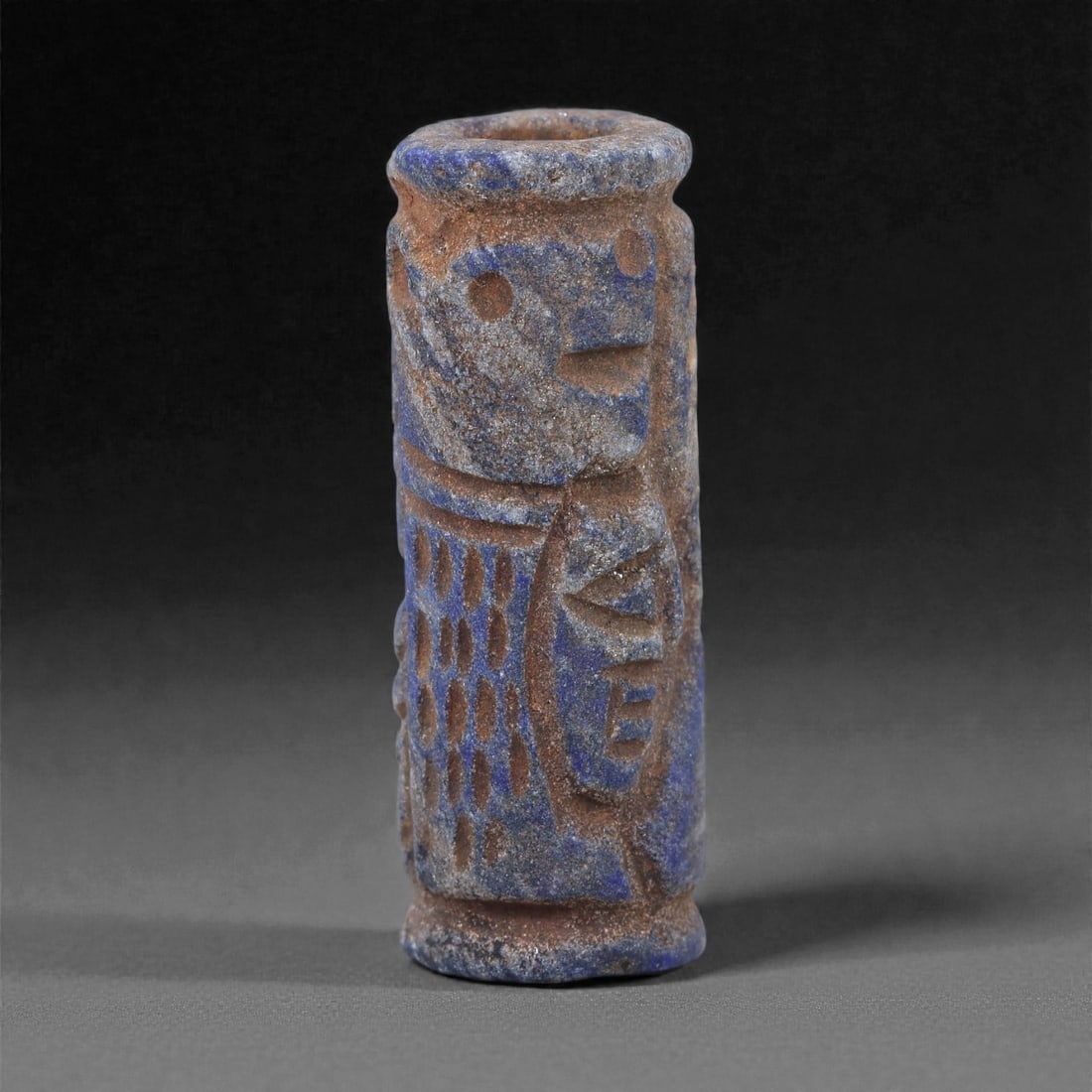 A Near Eastern Lapis Lazuli Cylindrical Relief Amulet, 3rd Millennium BCE, 3.4cm, 83L: Carved from lapis lazuli with mottled deep blue coloration interspersed with lighter calcite and brown mineral inclusions, the object is cylindrical in form with flat upper and lower faces. A longitud