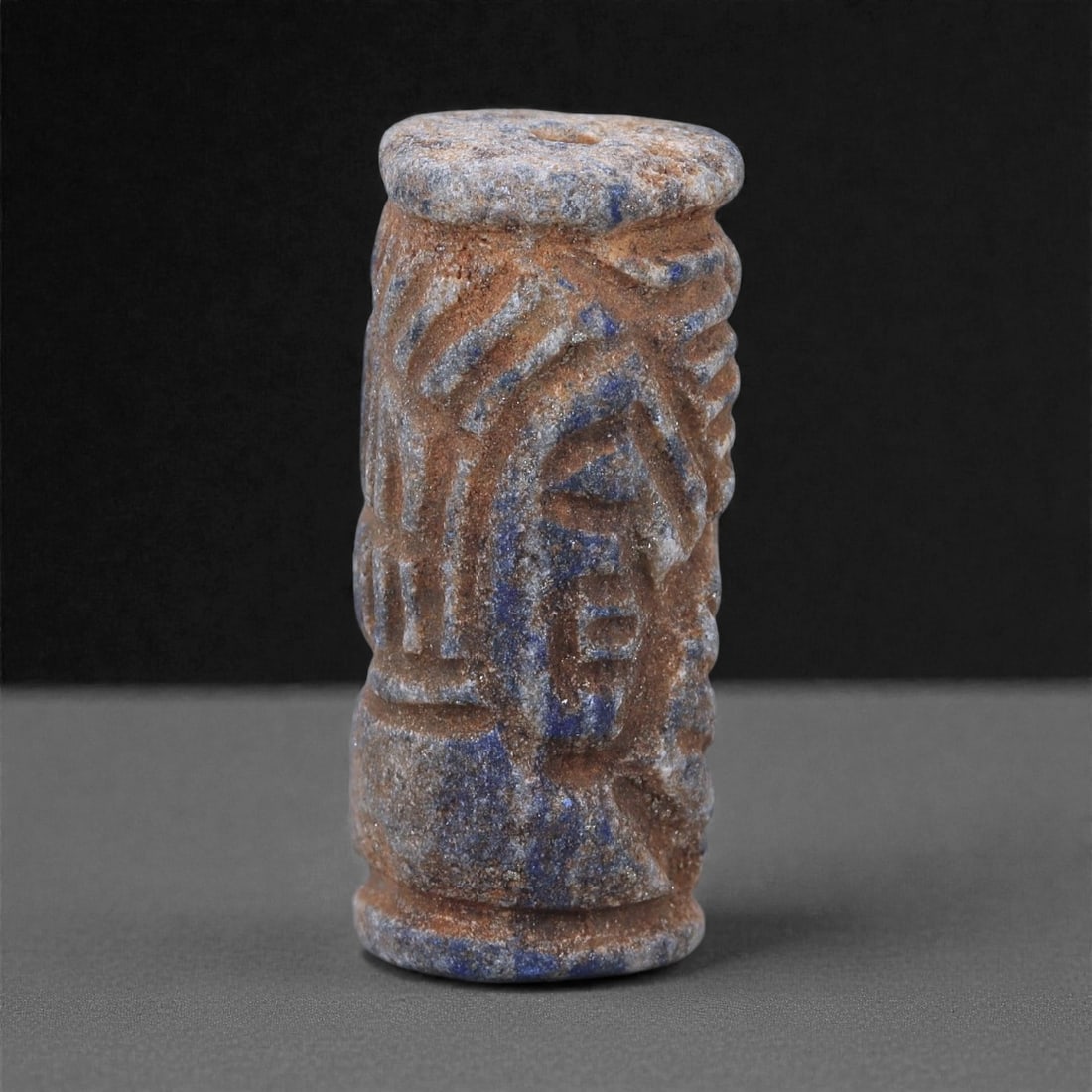 A Near Eastern Lapis Lazuli Cylindrical Relief Amulet, 3rd Millennium BCE, 2.7cm, 85L: Carved from lapis lazuli with mottled deep blue coloration interspersed with lighter calcite and brown mineral inclusions, the object is cylindrical in form with flat upper and lower faces. A longitud