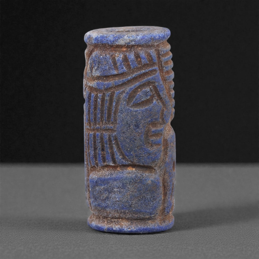 A Near Eastern Lapis Lazuli Cylindrical Relief Amulet, 3rd Millennium BCE, 3.6cm, 99L: Carved from lapis lazuli with mottled deep blue coloration interspersed with lighter calcite and brown mineral inclusions, the object is cylindrical in form with flat upper and lower faces. A longitud