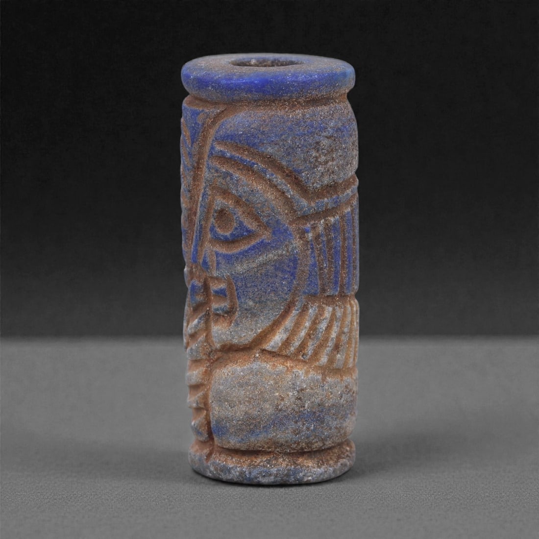 A Near Eastern Lapis Lazuli Cylindrical Relief Amulet, 3rd Millennium BCE, 3.5cm, 97L: Carved from lapis lazuli with mottled deep blue coloration interspersed with lighter calcite and brown mineral inclusions, the object is cylindrical in form with flat upper and lower faces. A longitud