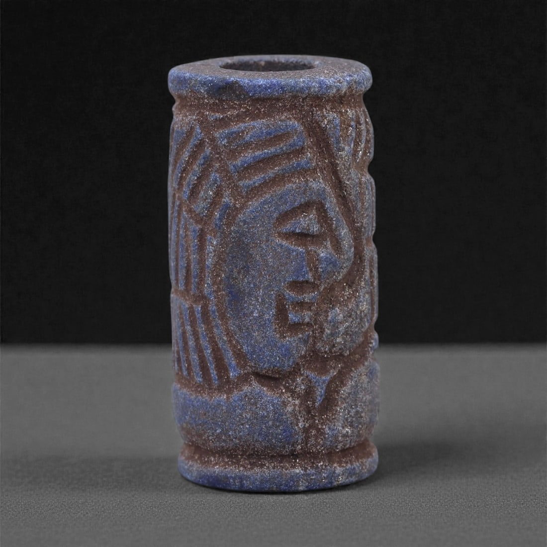 A Near Eastern Lapis Lazuli Cylindrical Relief Amulet, 3rd Millennium BCE, 2.7cm, 90L: Carved from lapis lazuli with mottled deep blue coloration interspersed with lighter calcite and brown mineral inclusions, the object is cylindrical in form with flat upper and lower faces. A longitud