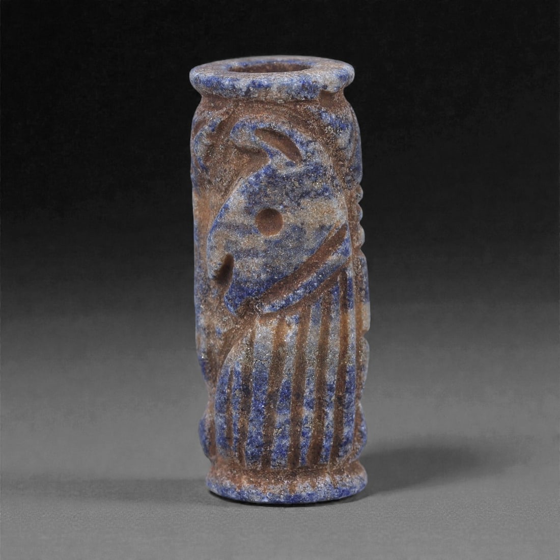 A Near Eastern Lapis Lazuli Cylindrical Relief Amulet, 3rd Millennium BCE, 3.3cm, 80L: Carved from lapis lazuli with mottled deep blue coloration interspersed with lighter calcite and brown mineral inclusions, the object is cylindrical in form with flat upper and lower faces. A longitud