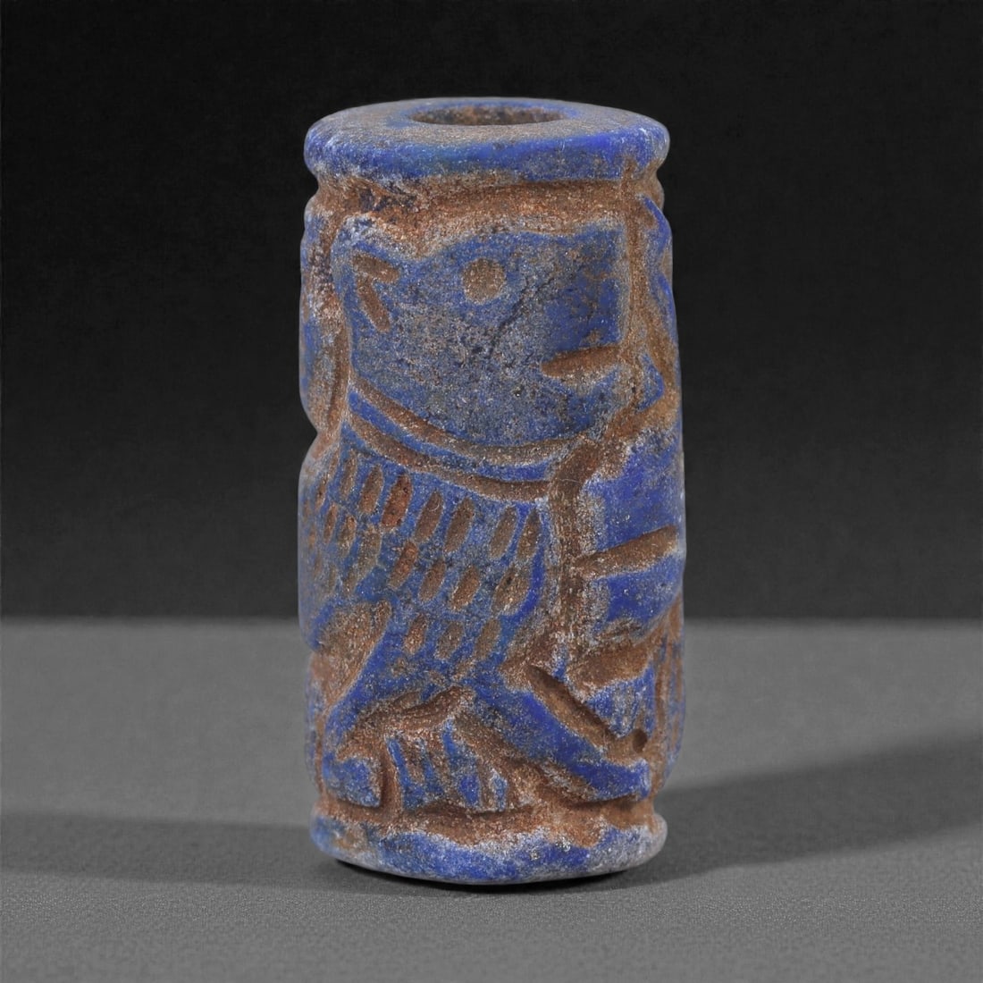 A Near Eastern Lapis Lazuli Cylindrical Relief Amulet, 3rd Millennium BCE, 3cm, 91L: Carved from lapis lazuli with mottled deep blue coloration interspersed with lighter calcite and brown mineral inclusions, the object is cylindrical in form with flat upper and lower faces. A longitud