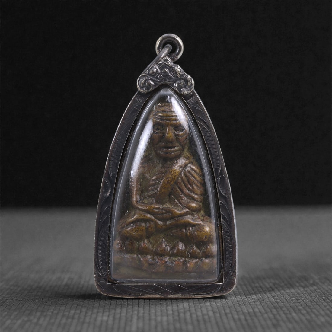 Thai Buddhist Monk Amulet - Seated Monk Pendant in Protective Frame, 29X: This devotional amulet features a seated Buddhist monk rendered in relief, housed within a fitted metal frame and clear protective cover. The monk is shown seated cross-legged on a stylized lotus base