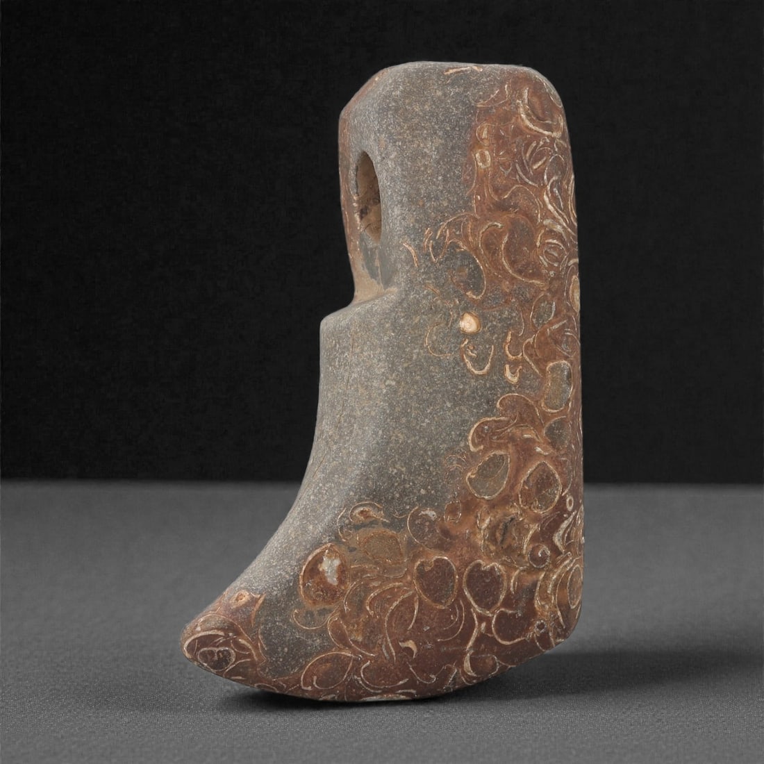 A Sino-Mongolian Fossilized Jasper Ritual Axe, 2nd-1st Millennium BCE, 65LR: Carved from fossil-bearing jasper with a mottled brown and grey surface, the object is shaped with a curved, downward-sweeping blade and a straight upper section rising into a perforated hafting ridge