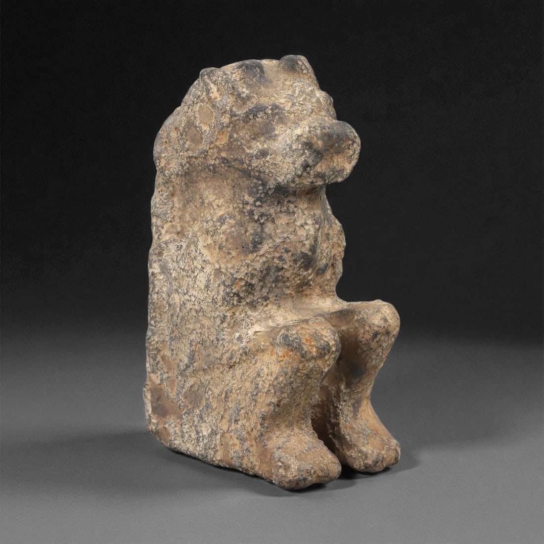An Egyptian Stone Lion Effigy, New Kingdom, 1550-1070 BCE, 65LE: Carved from a mottled beige and grey stone with surface weathering, the object represents a seated lion with the forelegs raised and bent at the knees. The head is rounded with a short projecting muzz
