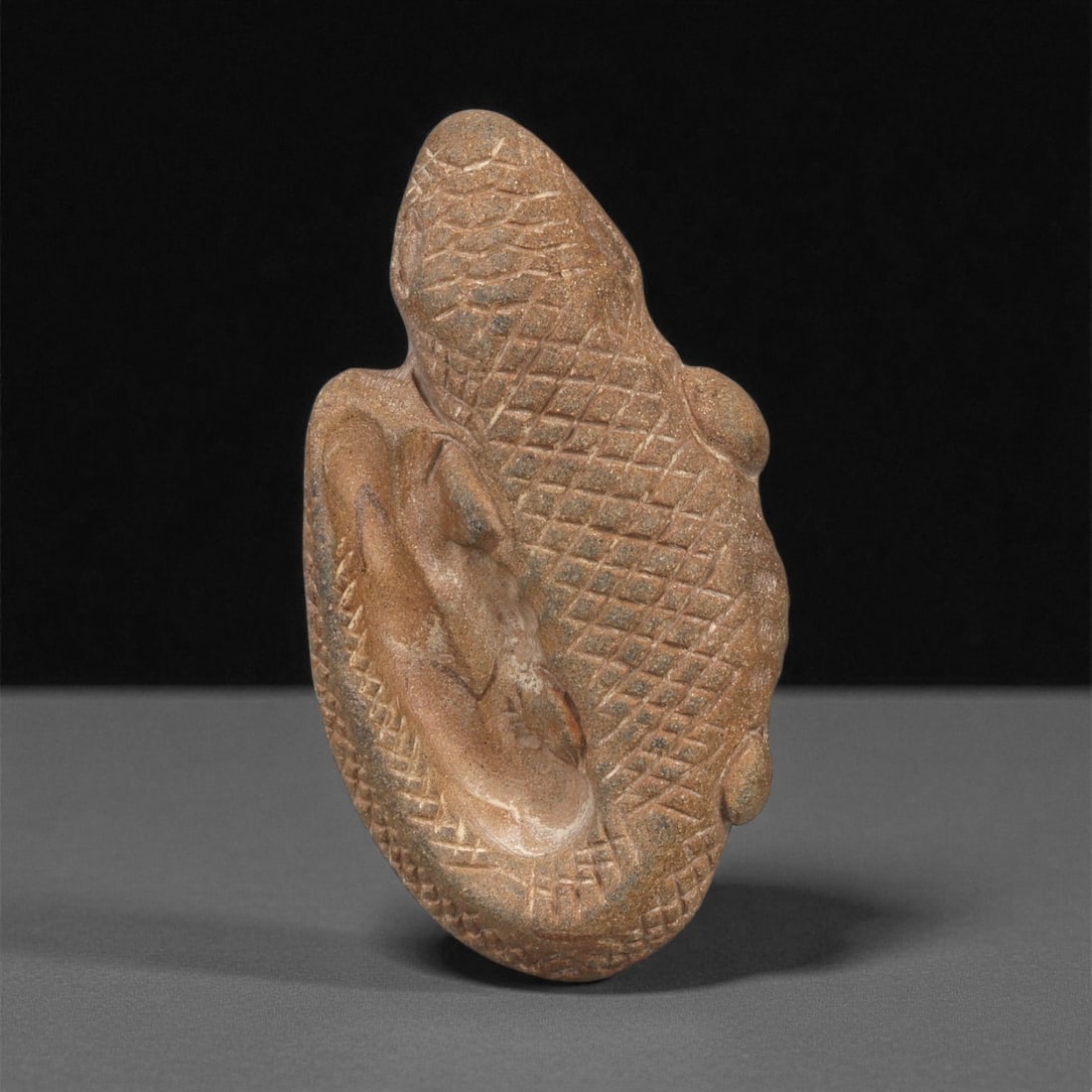 An Egyptian Stone Crocodile Figure, Predynastic Period, 4th Millennium BCE, 85ZB: Carved from a reddish-brown stone with a fine granular surface, the object represents a crocodile shown with its body tightly curved. The dorsal surface is covered with incised cross-hatched scales, w