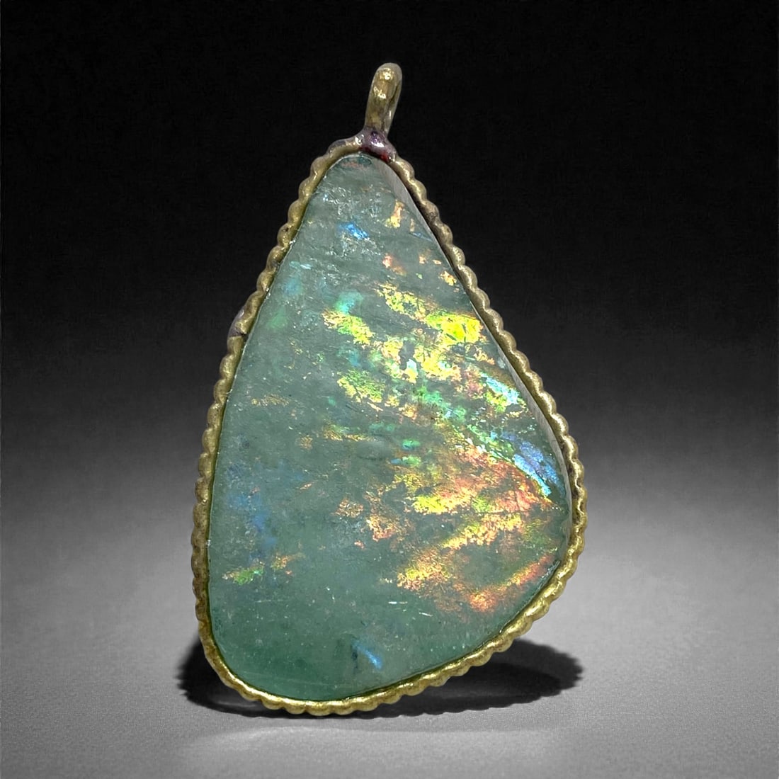 A Roman Glass Pendant, the Glass 1st-4th Century CE, Mounted in a Modern Setting, 70AA: Formed from a thin fragment of ancient Roman glass retaining a pale green body color beneath extensive iridescent weathering, the surface displays broad laminated bands of gold, blue, and rose tones t