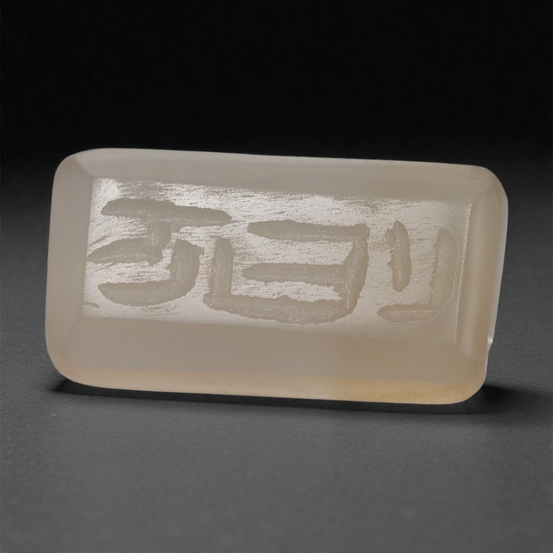 An Islamic Chalcedony Amuletic Plaque, Circa 9th-12th Century CE, 70A: Cut from pale translucent chalcedony, the plaque is rectangular in form with smoothly beveled edges. One face bears a shallowly engraved inscription executed in a cursive hand, the strokes rendered wi