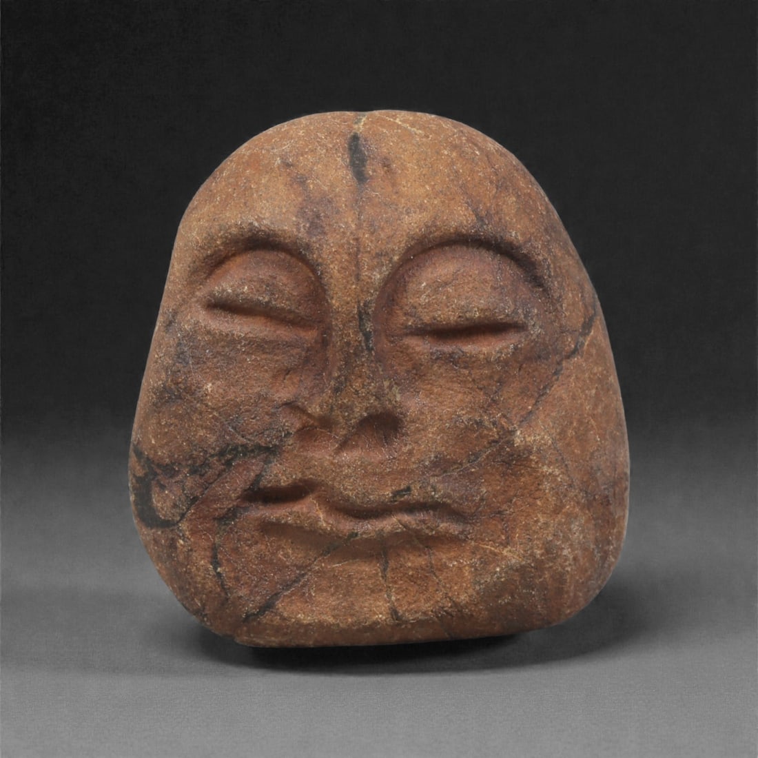 A Mediterranean Stone Maskette, 12th-17th Century, 44AC: Carved from a reddish-brown stone with pronounced dark veining, the maskette is formed with a broad upper section narrowing slightly toward the lower edge. The facial features are executed in shallow