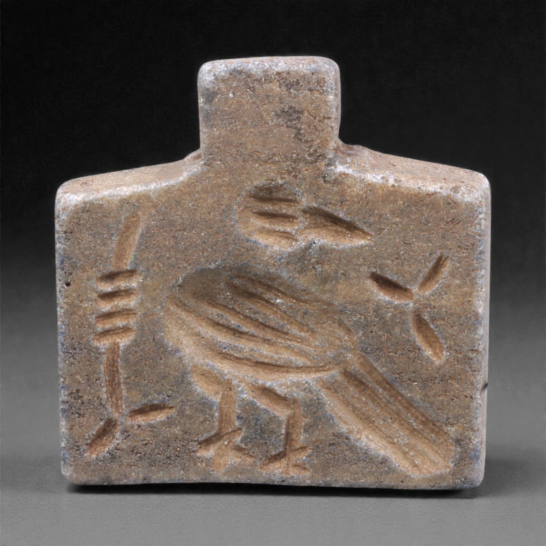 A Near Eastern Stone Bird Amulet, 3rd Millennium BCE, 15Q: Carved from a compact grey stone with areas of surface abrasion and mineral accretion, the amulet is formed as a small rectangular plaque with a stepped upper projection and double-convex faces. One f