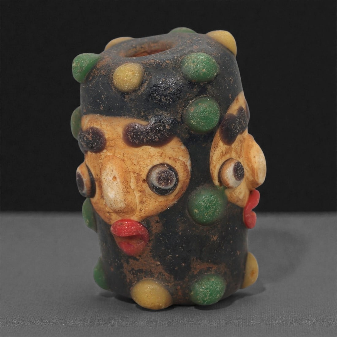 A European Glass Face Bead, 19th-20th Century, 69A: Formed in multicolored glass with an applied mask-like face, the bead is constructed around a cylindrical core with added elements in contrasting hues to create the eyes, brows, nose, and mouth. Addit