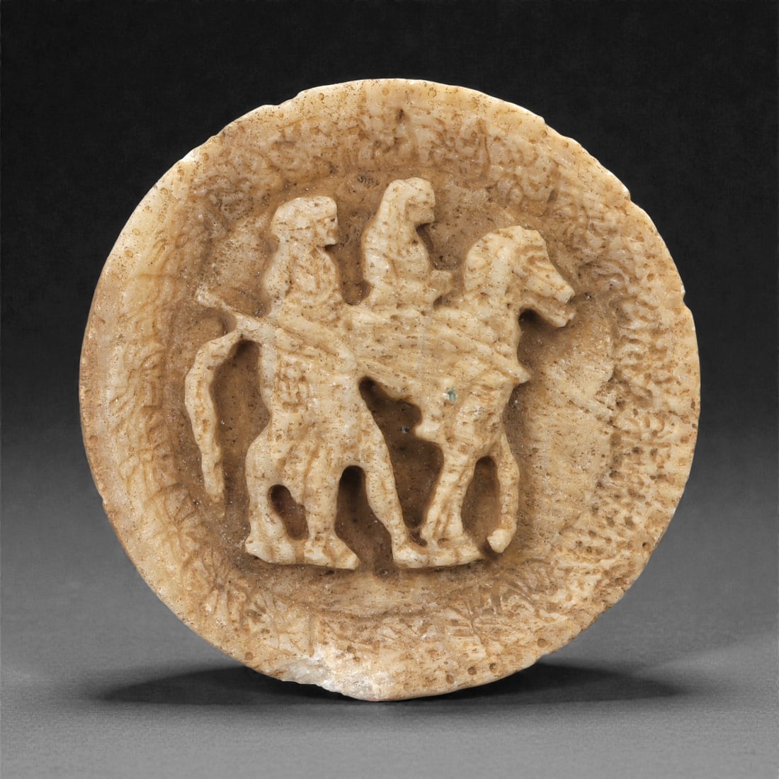 A Near Eastern Calcite Relief Plaque with Horse and Attendants, 1st Millennium CE, 15LN: Carved from pale calcite, the circular plaque features a low-relief scene showing a horse facing to the right, carrying a human rider with a second human figure positioned beside the animal. The forms