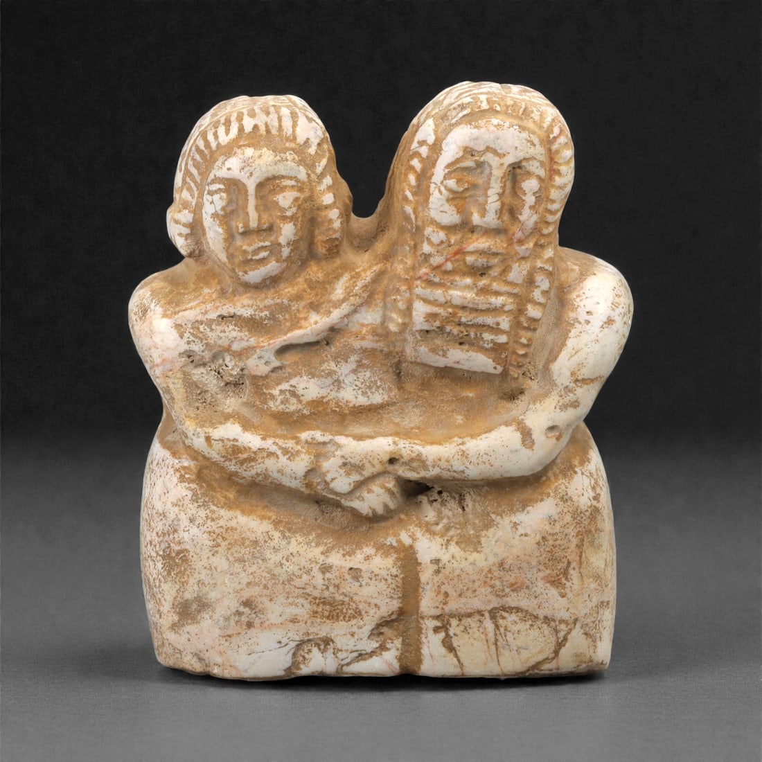 A Sumerian Stone Group Figurine Statue, 3rd Millennium BCE, 17LN: Carved from a light-colored stone with natural surface veining, the piece depicts two seated figures shown in close embrace, their arms rendered as broad, curving forms joining across the front. The h