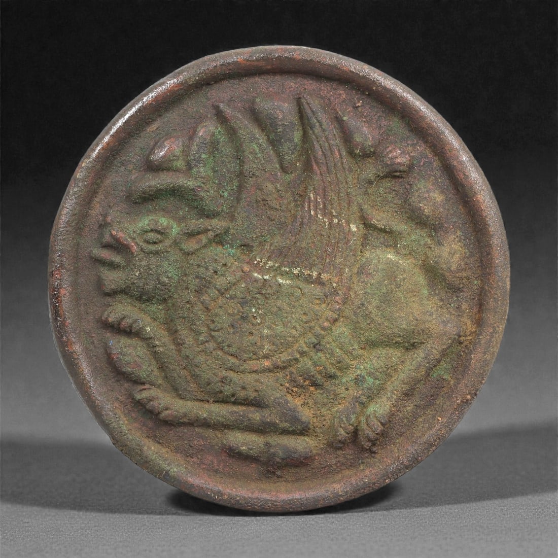 A Sasanian Bronze Stamp Seal with Winged Hybrid, 4th-7th Century CE, 68LP: Cast in bronze, the circular seal matrix is worked in recessed relief with a winged hybrid creature shown in profile. The figure displays a leonine body, a pronounced open muzzle, and a tall, backward