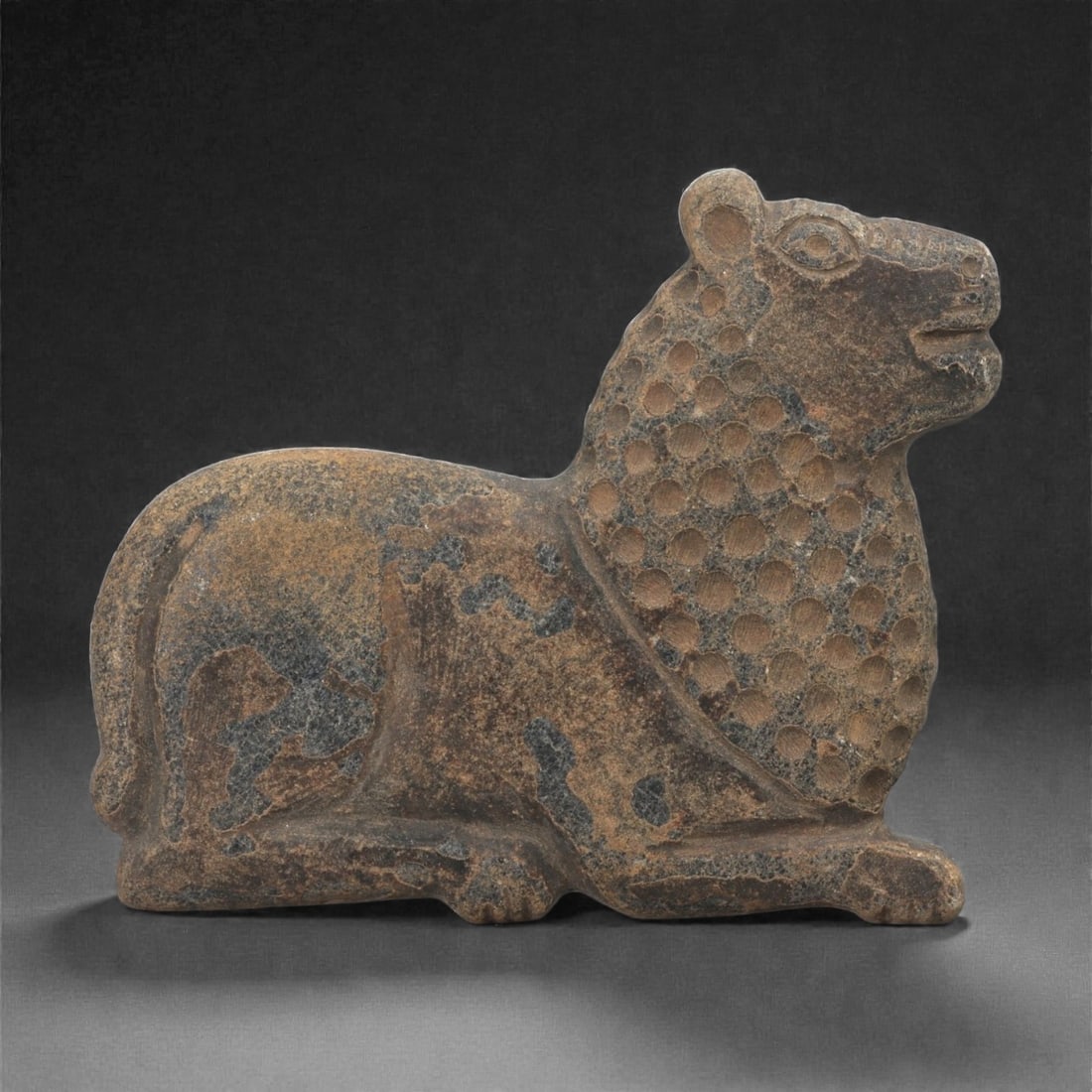 A Near Eastern Stone Feline Plaque, 1st Millennium BCE, 40LE: Carved from a brown stone with darker surface inclusions, the plaque depicts a recumbent feline in profile. The head is rendered with a rounded ear, defined eye, and open mouth, while the mane is indi