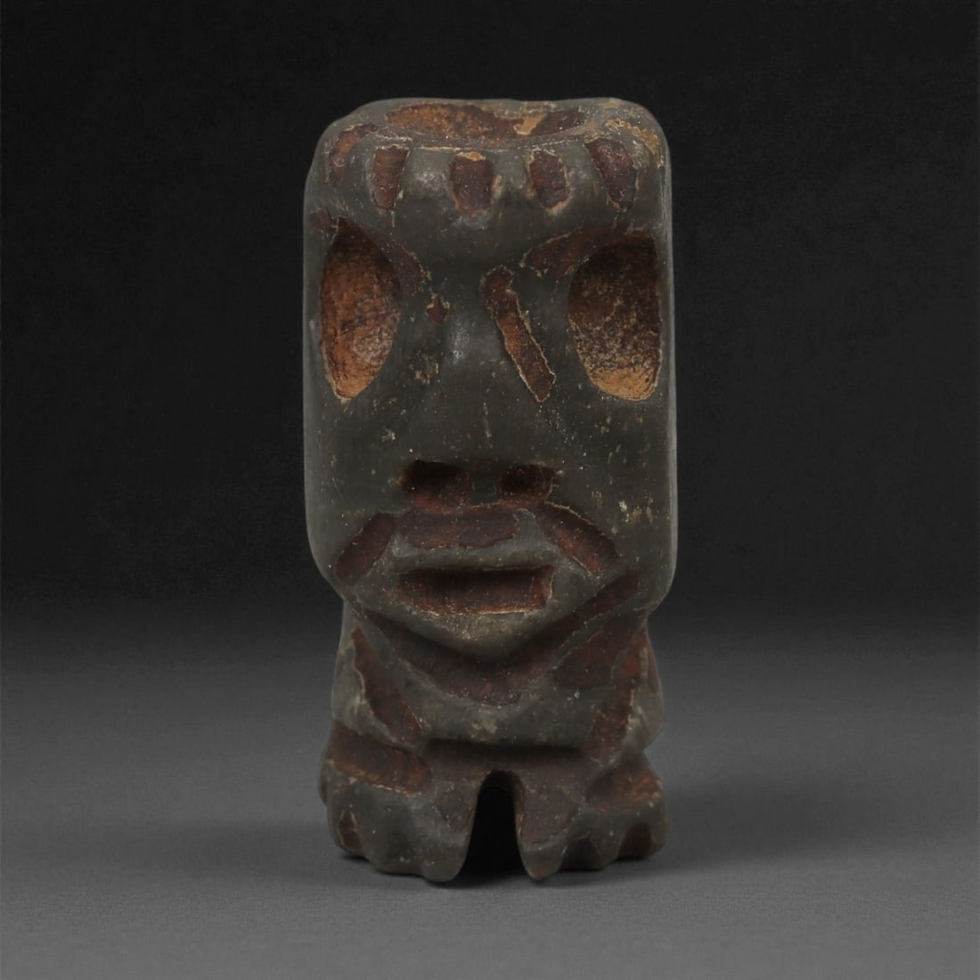 A Taino Carved Stone Idol, Late Pre-Columbian Period, Circa 1000-1500 CE, 92LA: Carved from a dark stone with reddish surface deposits, the figure is modeled with a squared head and compact lower body. The face bears recessed oval eyes, a central incised line along the nose, and
