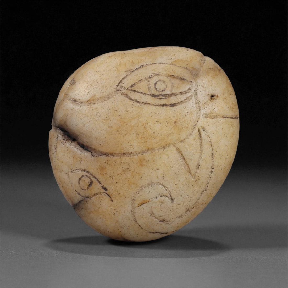An Egyptian Engraved Agate Amulet, New Kingdom-Late Period, c. 1550-332 BCE, 31LE: Carved from a pale beige agate with natural internal veining, the amulet is shaped as an irregular rounded pebble. Both sides bear incised linear motifs, including large almond-shaped eyes with centra
